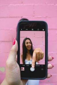 A woman captures her selfie using a smartphone against a vibrant pink wall.